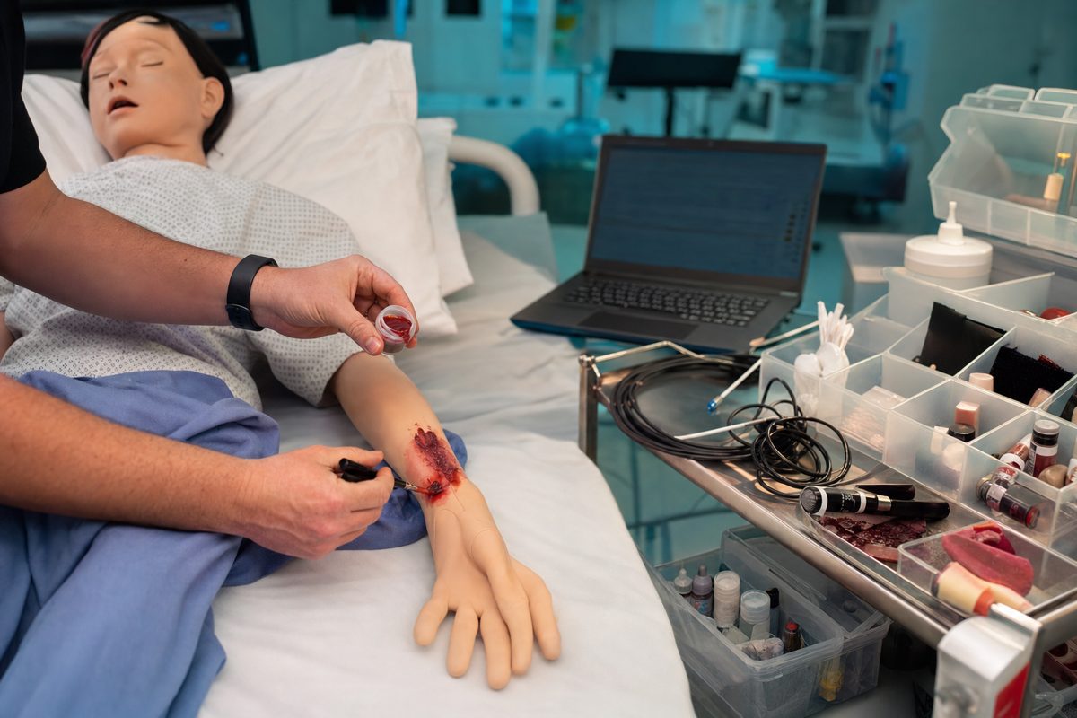 Hands applying moulage wound makeup to a training manikin, with a laptop, coiled cables, and supply bins on a prep cart